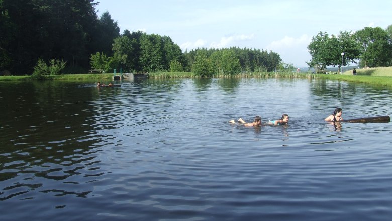 Naturbadeteich St. Martin, © Marktgemeinde St. Martin Menschen schwimmen in einem Naturbadeteich umgeben von Bäumen.
