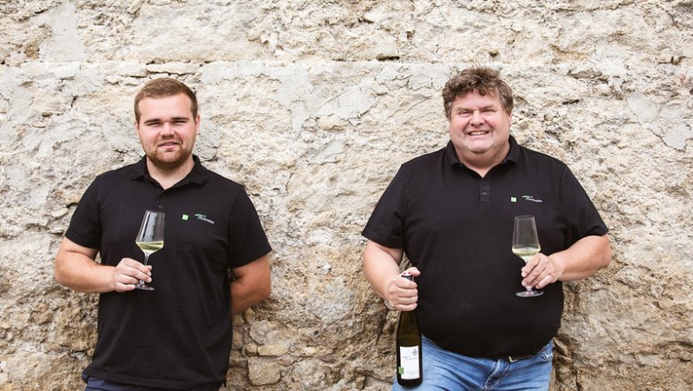 Meinhard und Daniel Forstreiter, © Weingut Meinhard Forstreiter Zwei Männer in schwarzen Polohemden mit Weingläsern vor einer Steinmauer.