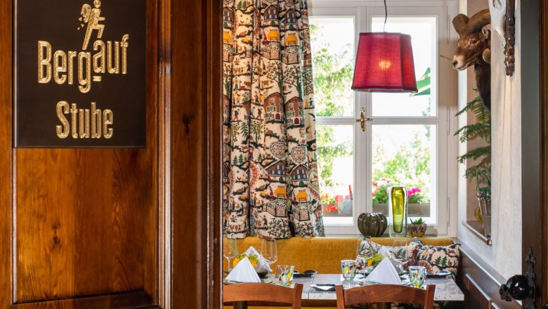 Restaurant Knappenhof, © Knappenhof - Christian Husar Ein gemütlicher Raum in einem Restaurant mit Holzmöbeln, einem Tisch mit gedecktem Geschirr, bunten Vorhängen und einem Geweih an der Wand.