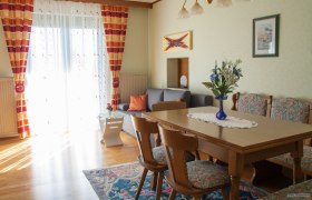 Wohnzimmer, © Fam. Sulzbacher Ein gemütliches Wohnzimmer mit Holztisch, Stühlen, grauem Sofa und bunten Vorhängen. Ein Blumenstrauß steht auf dem Tisch.