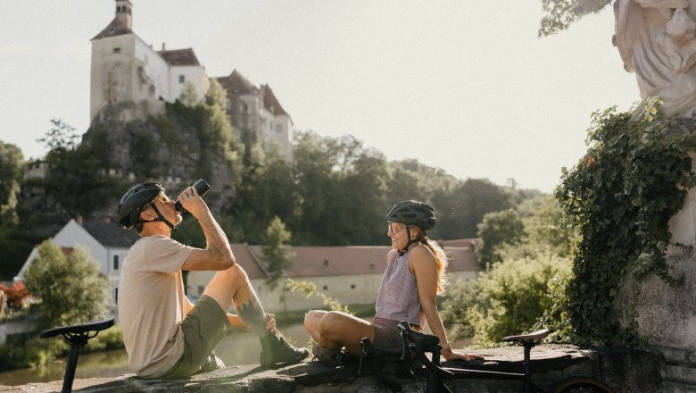 Burg Raabs, © Waldviertel Tourismus, Patrick Wasshuber Zwei Radfahrer mit Helmen sitzen auf einer Mauer vor der Burg Raabs.