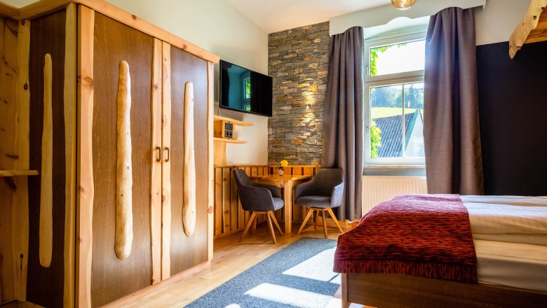 Land-Pension Kaiserhof, © Scharfegger´s Raxalpen Resort Gemütliches Zimmer mit Holzmöbeln, Steinwand, Tisch und Stühlen, Fernseher und Fenster mit Vorhängen.