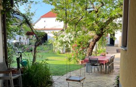 Herzlich Willkommen, © Gästehaus Weinwurm Ein idyllischer Garten mit einer Schaukel, einem Tisch mit Stühlen und blühenden Pflanzen.