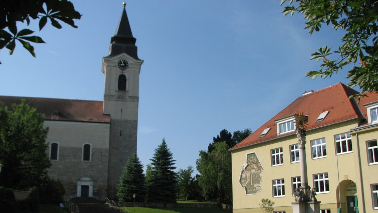 Stronsdorf, © Gemeinde Stronsdorf Kirche und Gebäude in Stronsdorf mit Uhrturm und Sonnenschein.