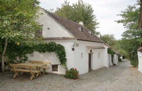 Weinviertler Kellergassenkulinarium, © D`Raschalaer Köllamauna Weißes Kellerstöckl mit Weinstöcken und Holztisch auf Kopfsteinpflasterstraße.