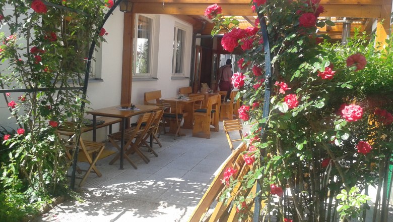 Rosenbogen, © Dominik Herzog Ein Rosenbogen mit roten Rosen führt zu einem Außenbereich mit Holztischen und -stühlen.