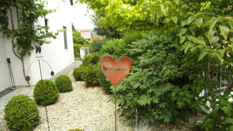 Landhaus Rossatz, © Familie Schlager Garten mit Herz-Willkommensschild und grünen Büschen vor einem weißen Gebäude.
