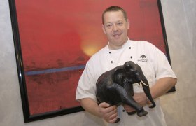 Restaurant zum schwarzen Elefanten Heinz Eisbacher, © diewerbetrommel.at Ein Koch in weißer Uniform hält eine schwarze Elefantenfigur vor einem roten Gemälde.