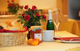 Haus Oestreicher, © Haus Oestreicher Frühstückstisch mit Brötchenkorb, Marmelade, Sektflasche und Rosen.