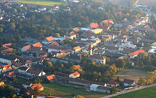 Münchendorf, © Wikipedia Luftaufnahme eines Dorfes mit Kirche und Wohnhäusern, umgeben von Bäumen und Feldern.