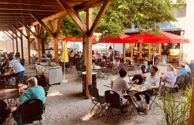 Brauhaus Schwechat, Gastgarten, © Brauhaus Schwechat Ein Biergarten mit Holzdach, roten Sonnenschirmen und vielen Menschen an Tischen.