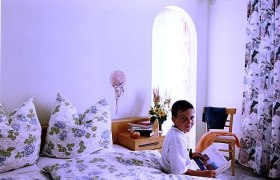 Gästezimmer, © Berg-Hahn Ein Junge sitzt auf einem Bett in einem hellen Zimmer mit floralen Vorhängen und Bettwäsche.