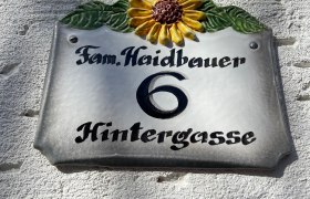 Pension Haidbauer, © Wiener Alpen Hausnummernschild mit Sonnenblume und der Aufschrift 'Fam. Haidbauer 6 Hintergasse'.