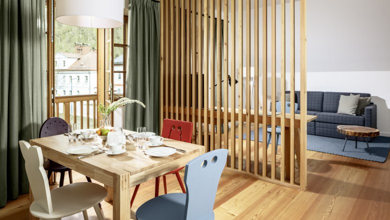 Hotel zum Glockenturm, © www.croce.at Ein helles Hotelzimmer mit Holzboden, Esstisch und Stühlen, abgetrennt durch eine Holzlamellenwand, mit Blick auf ein Sofa und einen Couchtisch.