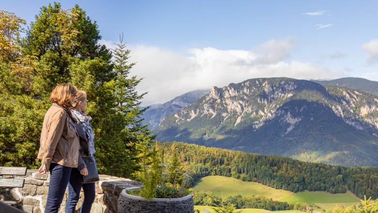 Beeindruckender Ausblick vom Looshaus, © Wiener Alpen / Christian Kremsl Zwei Personen stehen auf einer Terrasse und blicken auf eine Berglandschaft mit Wäldern und Wiesen.