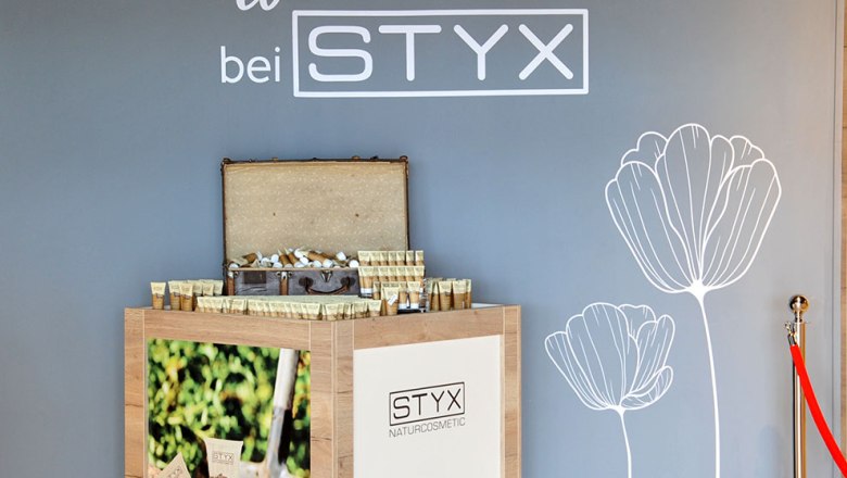 STYX Welcome Center- Shop, © Styx Naturkosmetic GmbH Ein Display mit STYX Naturkosmetik-Produkten vor einer grauen Wand mit Blumenzeichnung und Willkommensgruß.