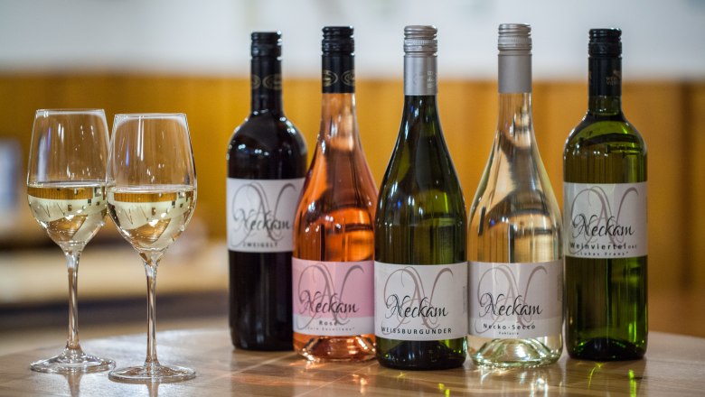 Neckam-Weine, © Schmeiser Fotographie Fünf Weinflaschen und zwei gefüllte Weingläser auf einem Holztisch.