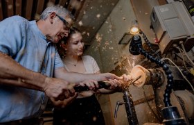 holzARTbinder - Kunstwerke aus Holz, © Waldviertel Tourismus, Matthias Streibel Zwei Personen arbeiten gemeinsam an einer Drehbank, um ein Holzstück zu bearbeiten. Holzspäne fliegen in der Luft.