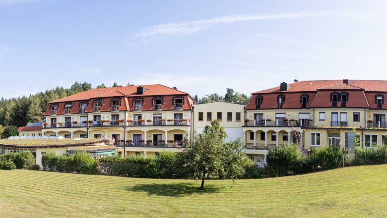 Außenansicht Gesundheitshotel Moorbad Bad Großpertholz, © Gesundheitshotel Moorbad Bad Großpertholz Außenansicht des Gesundheitshotels Moorbad Bad Großpertholz mit rotem Dach und Balkonen, umgeben von grüner Landschaft.