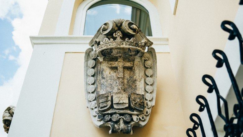 villa_schonthal-10_3, © Julius Hirtzberger Steinwappen an einer gelben Hauswand mit Fenster und blauer Himmel im Hintergrund.