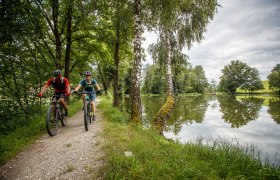 Granittrail, MTB, Mountainbiken, Radfahren, Yspertal, © Waldviertel Tourismus, Erwin Haiden Zwei Mountainbiker genießen die frische Luft und die malerische Landschaft entlang eines ruhigen Weges, der von hohen Bäumen gesäumt ist. Der glitzernde See spiegelt die Wolken und das satte Grün der Umgebung wider, während die beiden Radfahrer mit Freude und Energie in die Natur eintauchen.