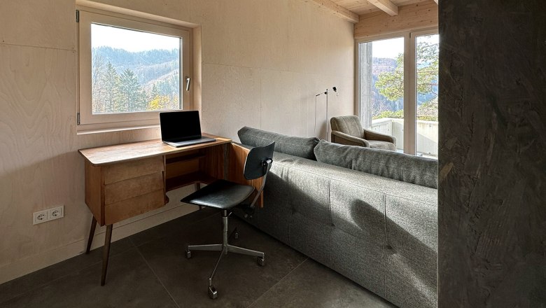 Arbeitsplatz, © Meinrad Hofer Ein moderner Arbeitsplatz mit einem Laptop auf einem Holztisch, einem schwarzen Stuhl und einem Sofa in einem Raum mit großen Fenstern und Waldblick.