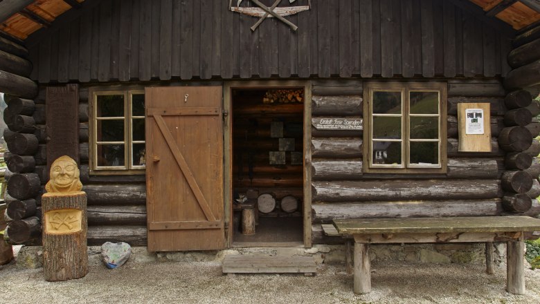 Holzknechthütte, © Wiener Alpen, Foto: Bene Croy Eine rustikale Holzhütte mit offener Tür, Holzschnitzerei und Tisch davor.