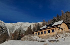 Waxriegelhaus, © Martin Tod - Waxriegelhaus Ein Holzhaus im Schnee vor einer Bergkulisse unter klarem, blauem Himmel.