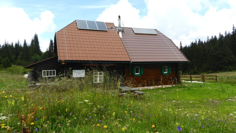 Huette2, © Richard Waidhofer Eine Berghütte mit Solarpanelen auf dem Dach, umgeben von einer blühenden Wiese und Wald im Hintergrund.