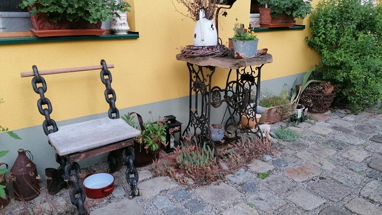 Upcycling Garten, © Tina Perina Ein Garten mit einer gelben Wand, einem Stuhl aus Ketten und Holz, und einem Tisch aus einer alten Nähmaschine, dekoriert mit Pflanzen.