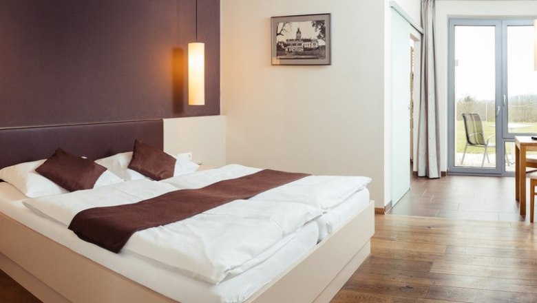 Ruhige Zimmer mit großem Komfort., © Weingut & Weinblick Grafinger Modern eingerichtetes Hotelzimmer mit Doppelbett, Holzboden und Zugang zu einem Balkon mit Ausblick.