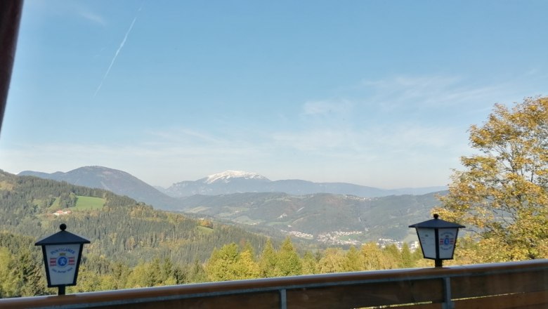 Aussicht aus dem Restaurant, © Wiener Alpen Blick von einem Restaurantbalkon auf eine bergige Landschaft mit Wald und einem schneebedeckten Gipfel im Hintergrund.
