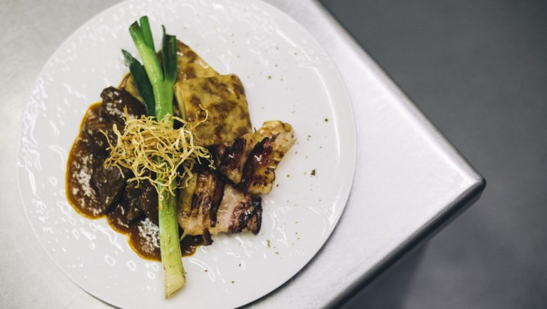 Rostbraten von der Alpenvorland Beiried, © Niederösterreich Werbung/David Schreiber Ein Teller mit Rostbraten, Kartoffelgratin, Lauch und knusprigen Zwiebeln auf einer Metalloberfläche.