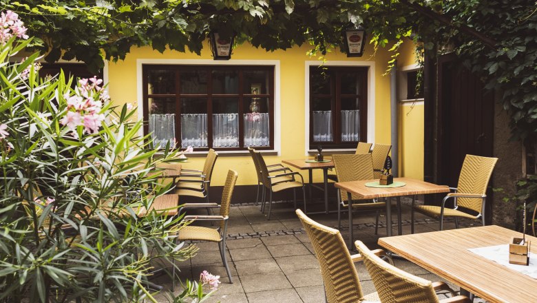 Ein typisch Weinviertler Wirtshaus eben, © Niederösterreich Werbung/David Schreiber Gemütlicher Gastgarten eines Wirtshauses mit gelber Fassade, Holztischen und Korbstühlen, umgeben von Pflanzen und Weinreben.