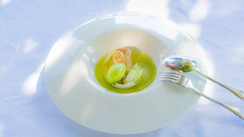 Kulinarische Genüsse im Hotel Richard Löwenherz, © Chris Saupper Ein elegantes Gericht auf einem weißen Teller mit grüner Sauce, einer Jakobsmuschel und grüner Garnitur, auf einem weißen Tischtuch serviert.