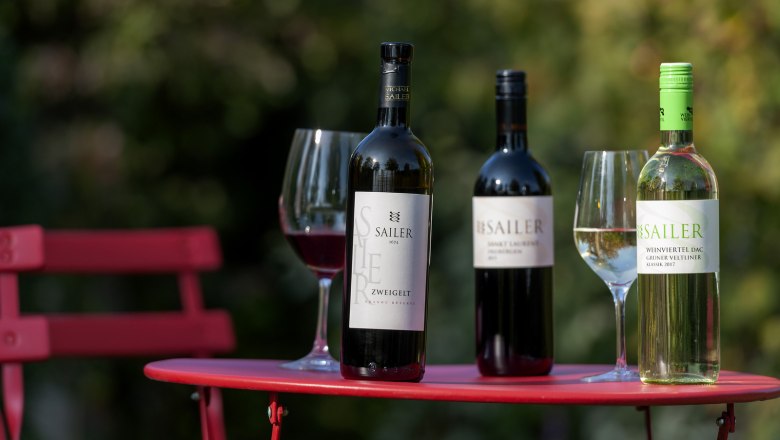 Weingut Michael Sailer, © Michael Himml Drei Weinflaschen und zwei Weingläser auf einem roten Tisch im Freien.