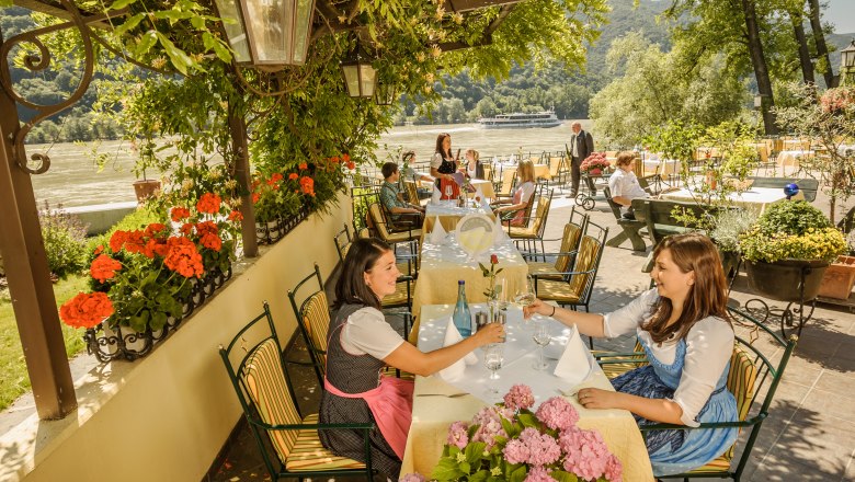 Garten Restaurant Heinzle, © Rita Newman Menschen sitzen in einem Gartenrestaurant mit Blick auf einen Fluss, umgeben von Blumen und Bäumen.