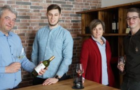Familie Mösslinger, © Weinbau Mösslinger Vier Personen stehen an einem Tisch mit Weingläsern und einer Weinflasche vor einer Ziegelwand.