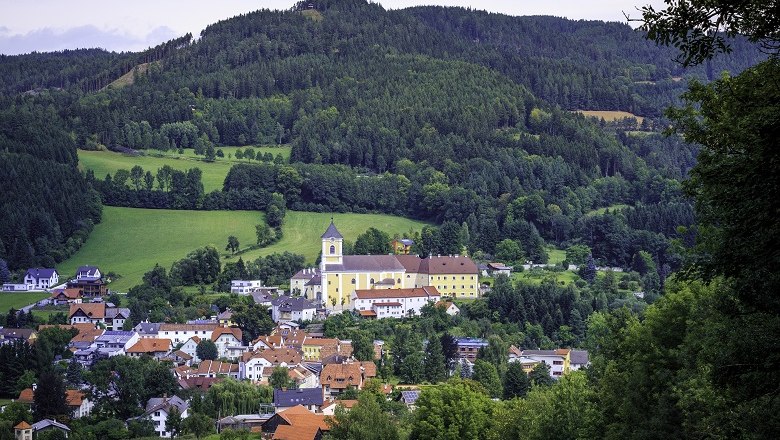 Imposant thront die Kirche über dem Ort, © Wiener Alpen, Kremsl Eine Kirche thront über einem malerischen Dorf, umgeben von grünen Hügeln und Wäldern.