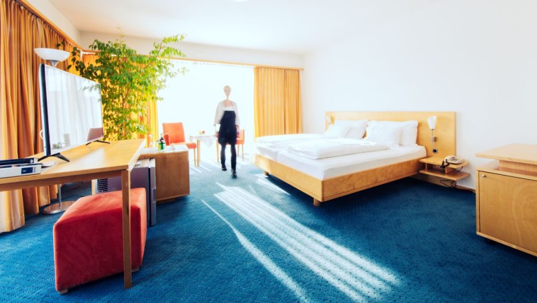 Wohnraum_3, © Franz Klinglhuber GmbH Helles Hotelzimmer mit Doppelbett, Schreibtisch, Fernseher und großer Pflanze. Eine Person geht durch den Raum.