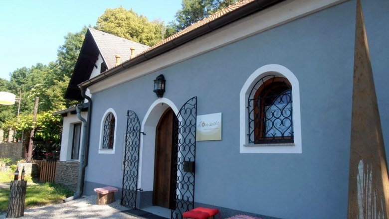 Loessiade, © Lössiade Ein kleines, blaues Gebäude mit Holztür und dekorativen Gittern vor den Fenstern. Davor stehen Holzhocker mit roten Kissen.