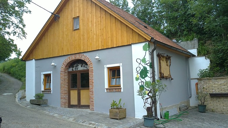Heurigenlokal, © Weinbau Fitz Ein traditionelles Heurigenhaus mit Holzverkleidung und Ziegelbogen, umgeben von Pflanzen.