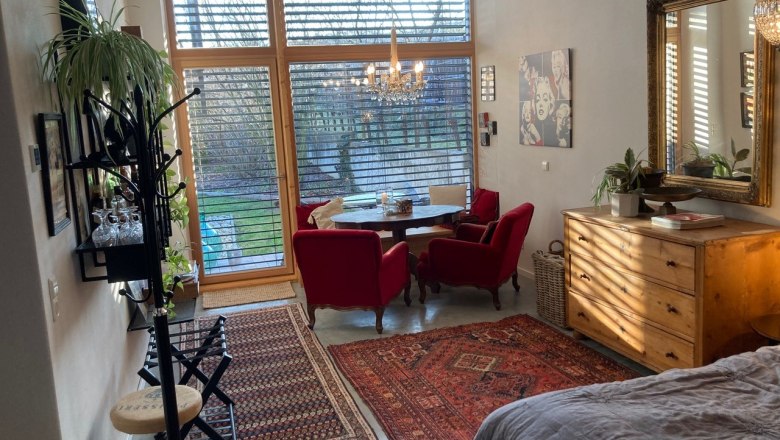 Apartment mit Blick in den Garten, © Weitschacher Wohnzimmer mit großen Fenstern, roten Sesseln, Holzmöbeln und Teppichen, Blick in den Garten.