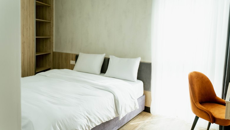 Hotel Lamede, © Hotel Lamede Modernes Hotelzimmer mit Doppelbett, weißen Bettlaken, Holzregal und orangefarbenem Stuhl vor einem Fenster mit Vorhängen.