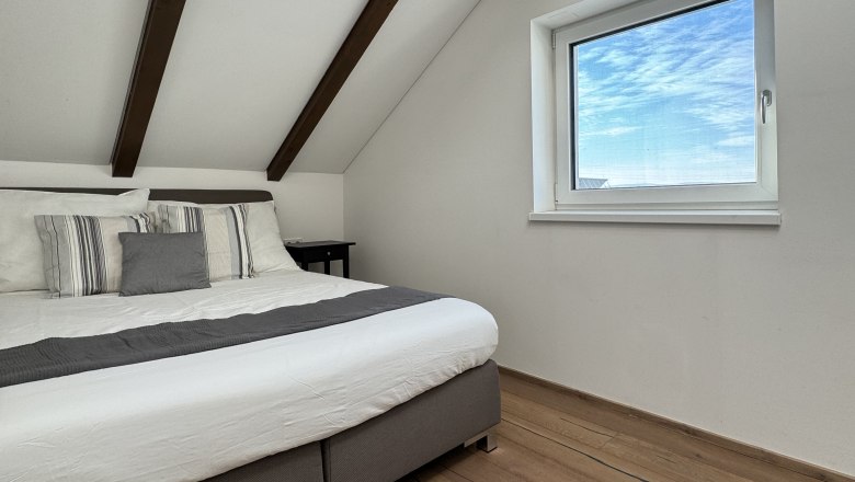 Schlafzimmer, © Juwel am Stadtrand Modernes Schlafzimmer mit Holzbalkendecke, großem Bett und Fenster mit Blick auf blauen Himmel.