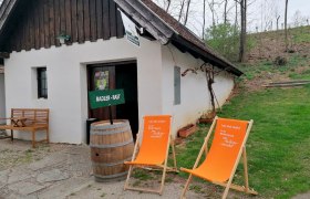 Radlerrast von Familie Holzschuh, © Martha Holzschuh Kleines weißes Gebäude mit Holzdach, zwei orange Liegestühle und ein Holzfass davor, umgeben von Bäumen und Gras.