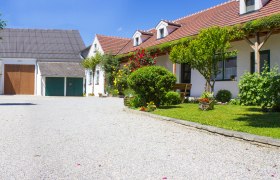 Urlaubsweingut, © Fam. Lobner Ein idyllisches Weingut mit Kiesweg, blühenden Gärten und traditionellen Gebäuden unter blauem Himmel.