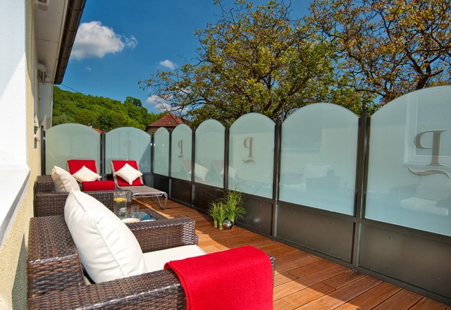 Wellnessterrasse, © Residenz Wachau Eine Terrasse mit Rattanmöbeln, roten Decken und Kissen, umgeben von Glaswänden und Bäumen im Hintergrund.