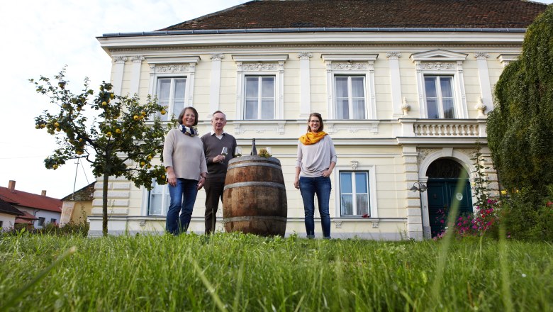 Familie Rücker, © Katharina F-Roßboth Drei Personen stehen vor einem großen, gelben Gebäude mit einem Fass im Vordergrund.