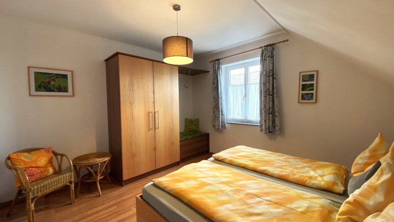 Ferienwohnung Marille, © Trachsler Schlafzimmer mit Doppelbett, Holzschrank, Korbstuhl und Fenster mit Vorhängen.